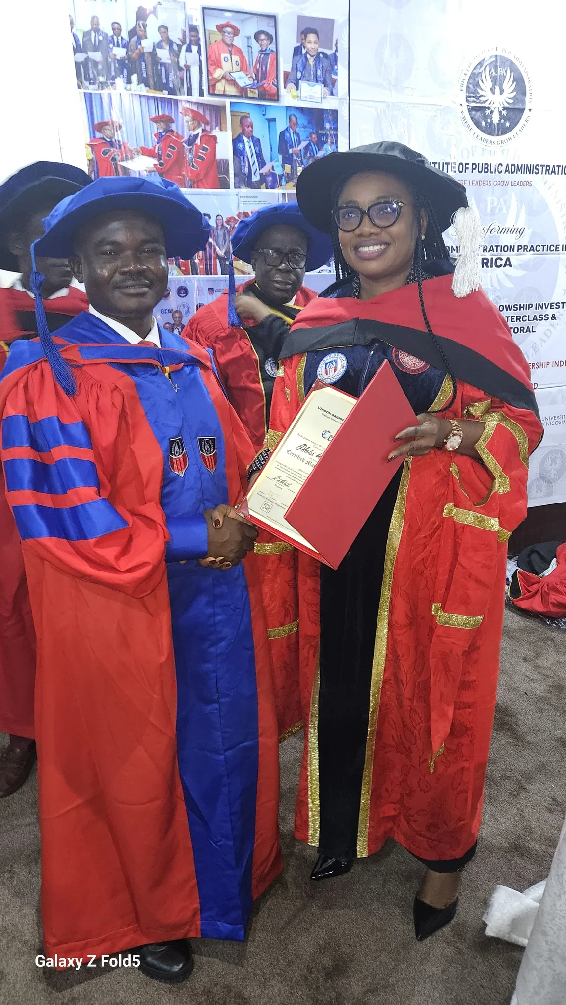 Dr. Alabi in a red and blue academic gown shaking hands and receiving a certificate from a senior academic administrator in red regalia.