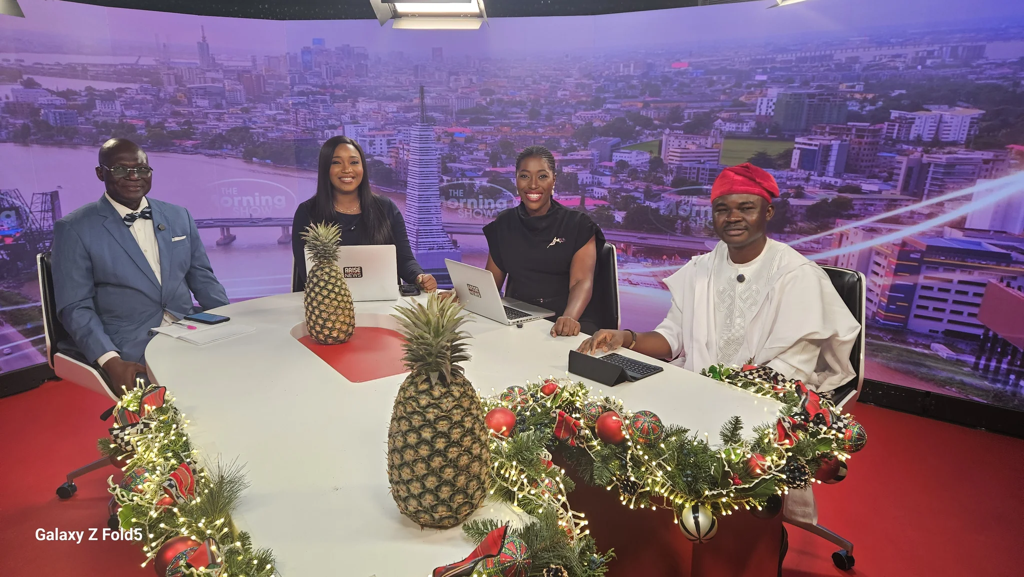 Wide studio camera shot of Dr. Alabi seated at the Arise News "The Morning Show" broadcast table with co-anchors and pineapples displayed as centerpieces.