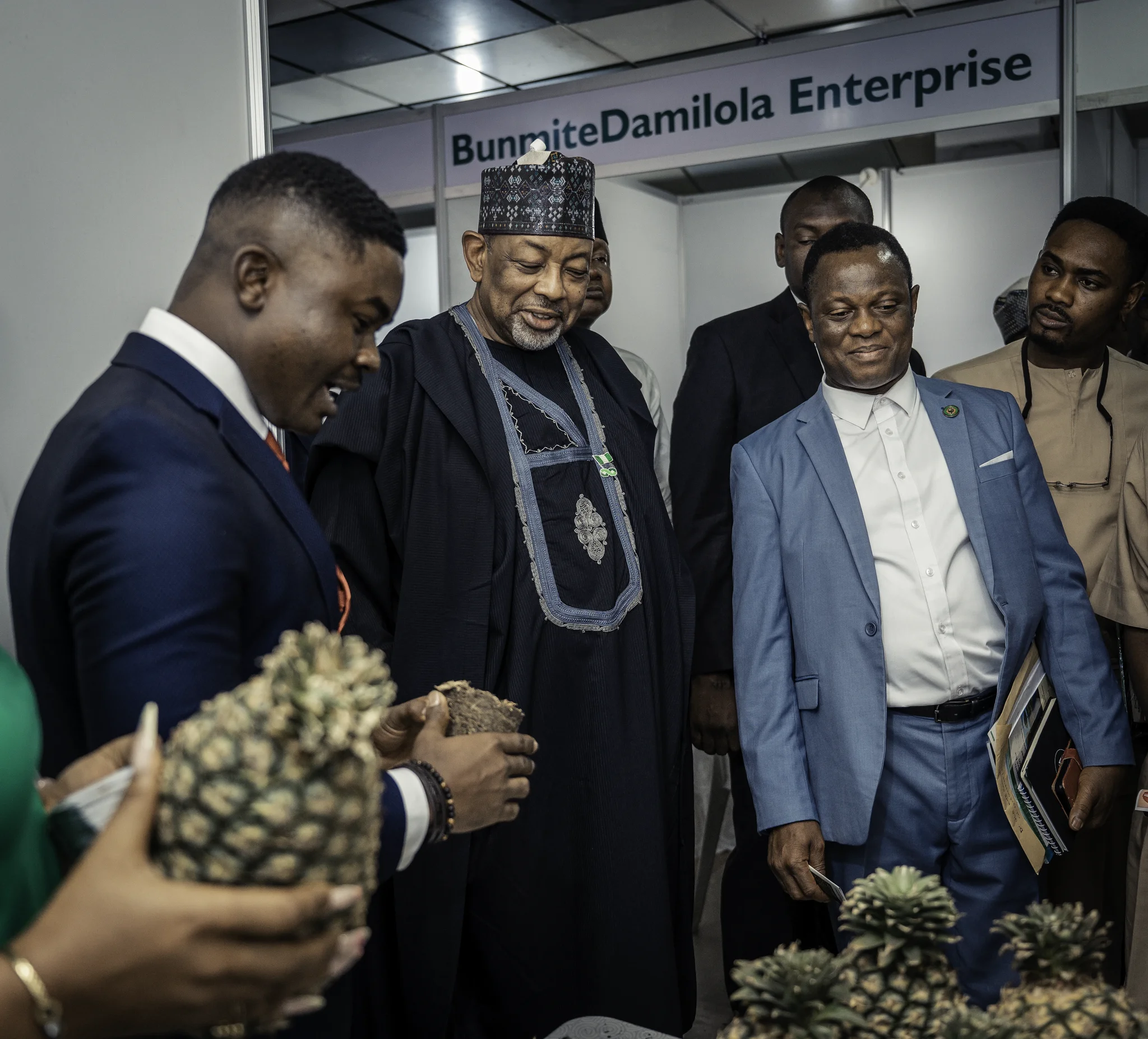 Dr. Alabi actively pointing and explaining value-added pineapple products to Minister Kyari and attending policy executives at a trade booth.