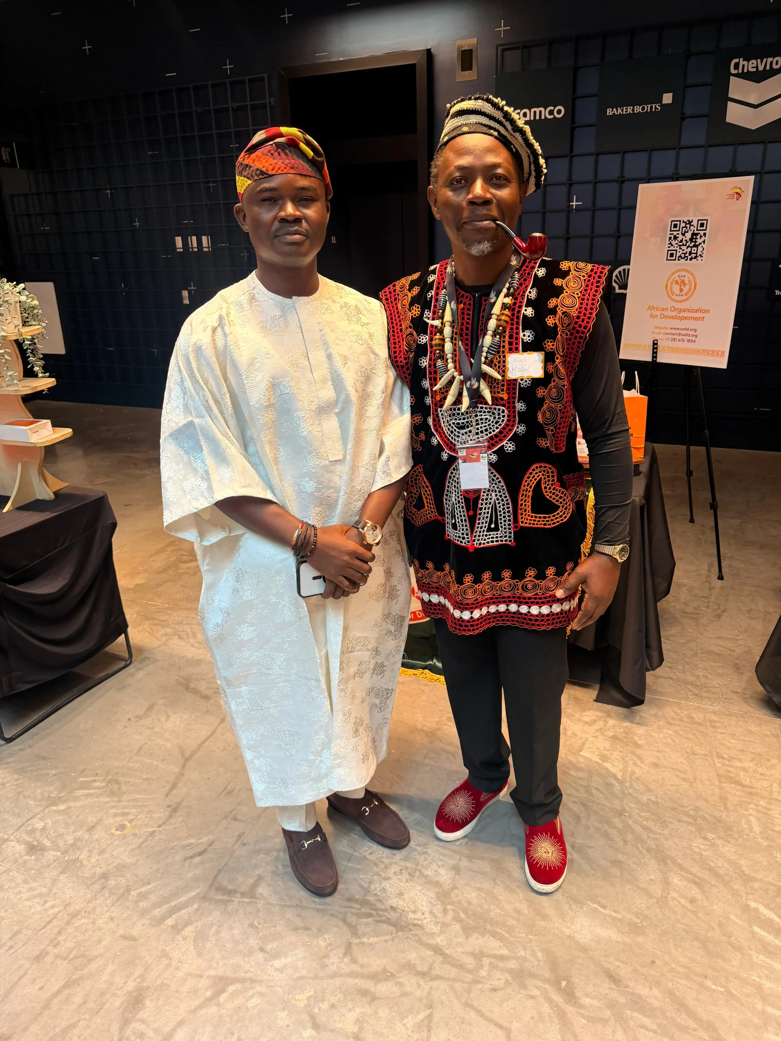 Full-length photo of Dr. Alabi in a white Agbada standing alongside an African cultural delegate in traditional ceremonial attire and smoking pipe at a Houston convention venue.