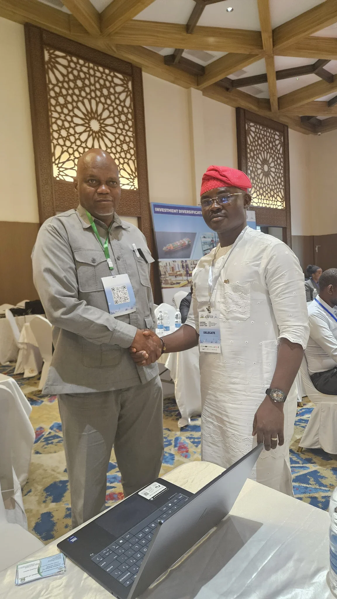 Dr. Segun Alabi in a white embroidered Agbada and red cap shaking hands with a summit delegate wearing a grey suit in a conference hall.