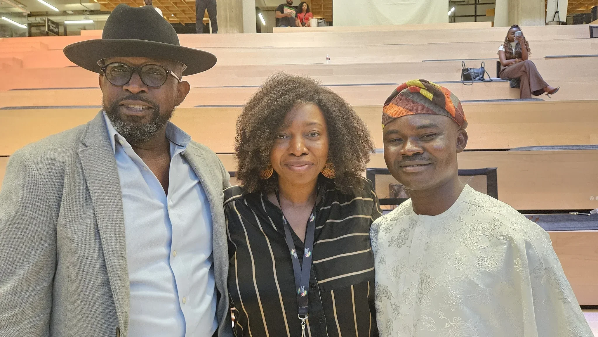 Dr. Alabi in a white Agbada posing warmly with a male executive in a grey blazer/hat and a female delegate inside the tiered amphitheater seating at the Diaspora Africa Conference.