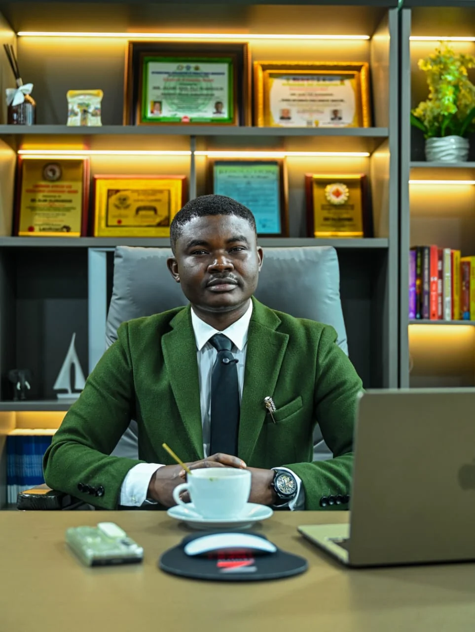 Perfectly composed studio-quality executive portrait of Dr. Alabi in a green blazer, seated behind his desk with a coffee cup, laptop, and awards displayed behind him.