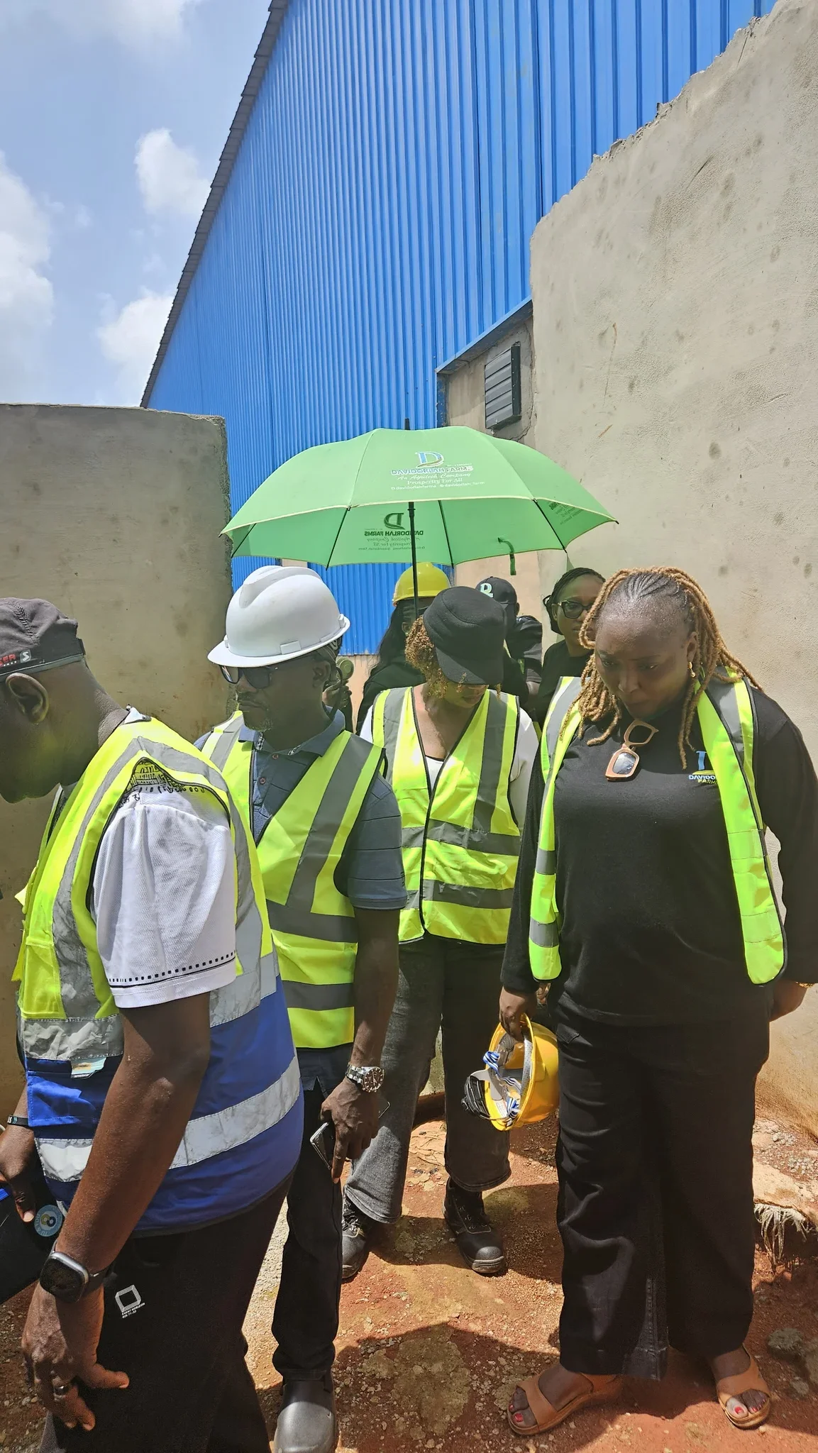 Operational inspection photo of engineers and executives in safety vests and hard hats holding a branded Davidorlah Farms umbrella outside a processing building.