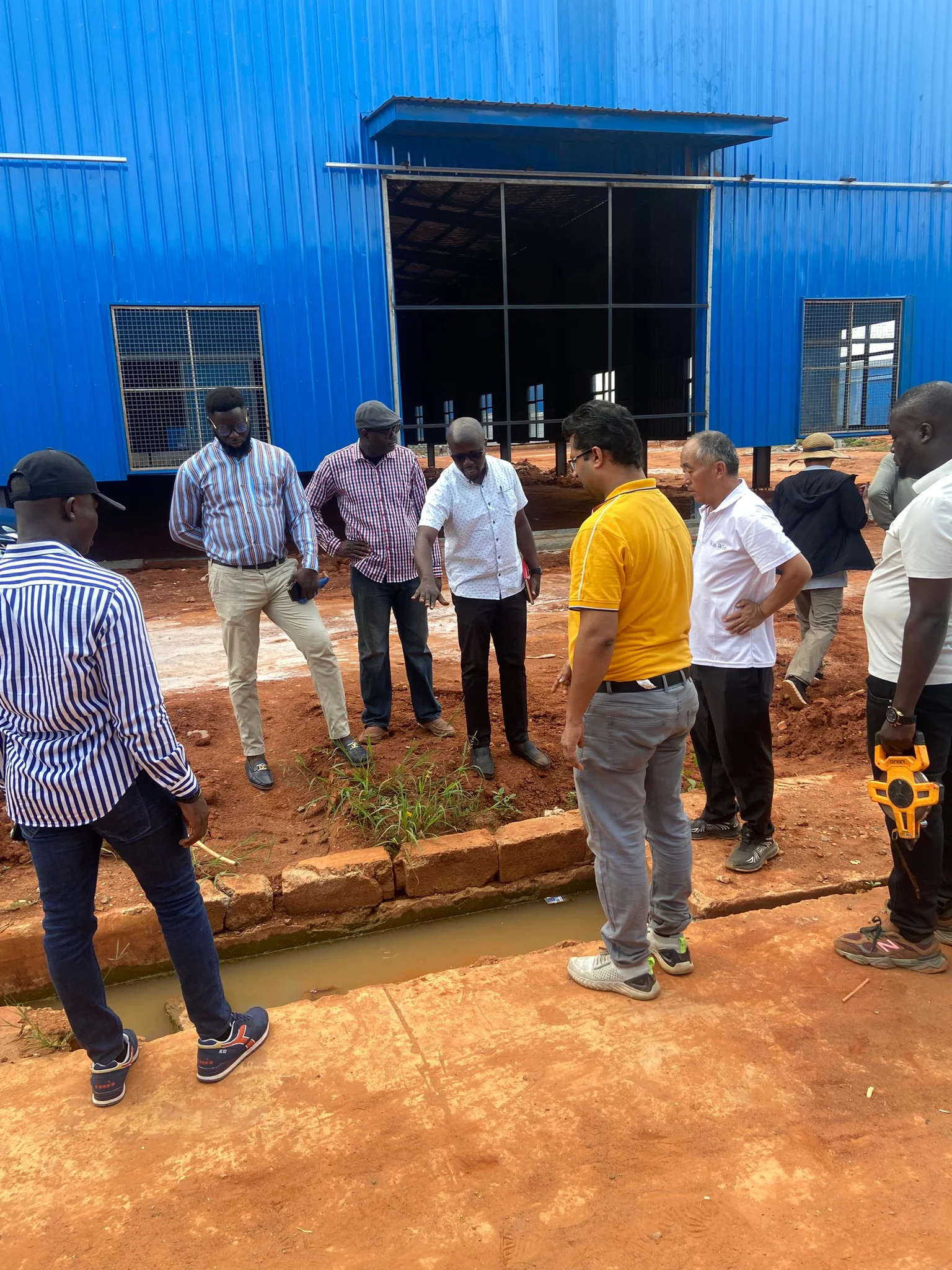 High-angle outdoor shot of an engineering and technical survey team standing in front of a blue industrial warehouse structure examining drainage and soil foundations.