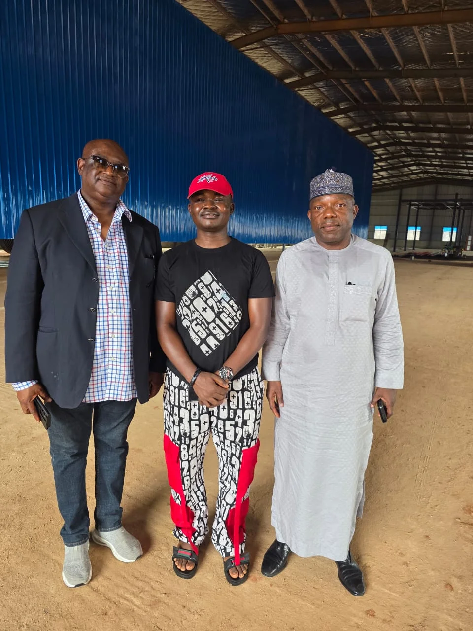 Dr. Alabi in a black t-shirt and red cap standing inside a large industrial processing warehouse alongside two corporate executives.