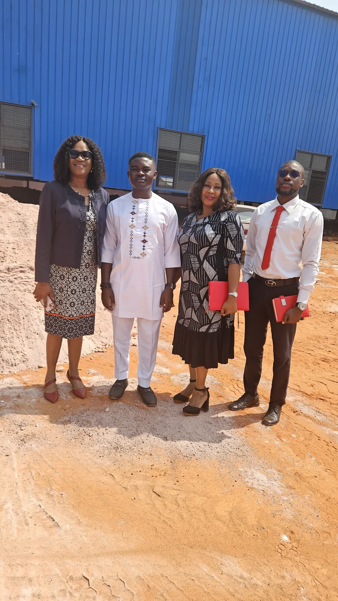 Dr. Alabi in a white tunic standing with a delegation of three corporate executives outside a large blue industrial processing structure.