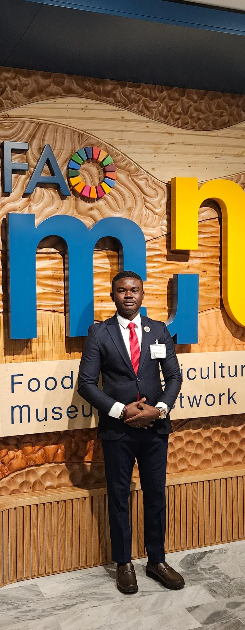 Full-length portrait of Dr. Alabi standing in front of the carved wooden UN FAO Food Museum Network installation in Rome.