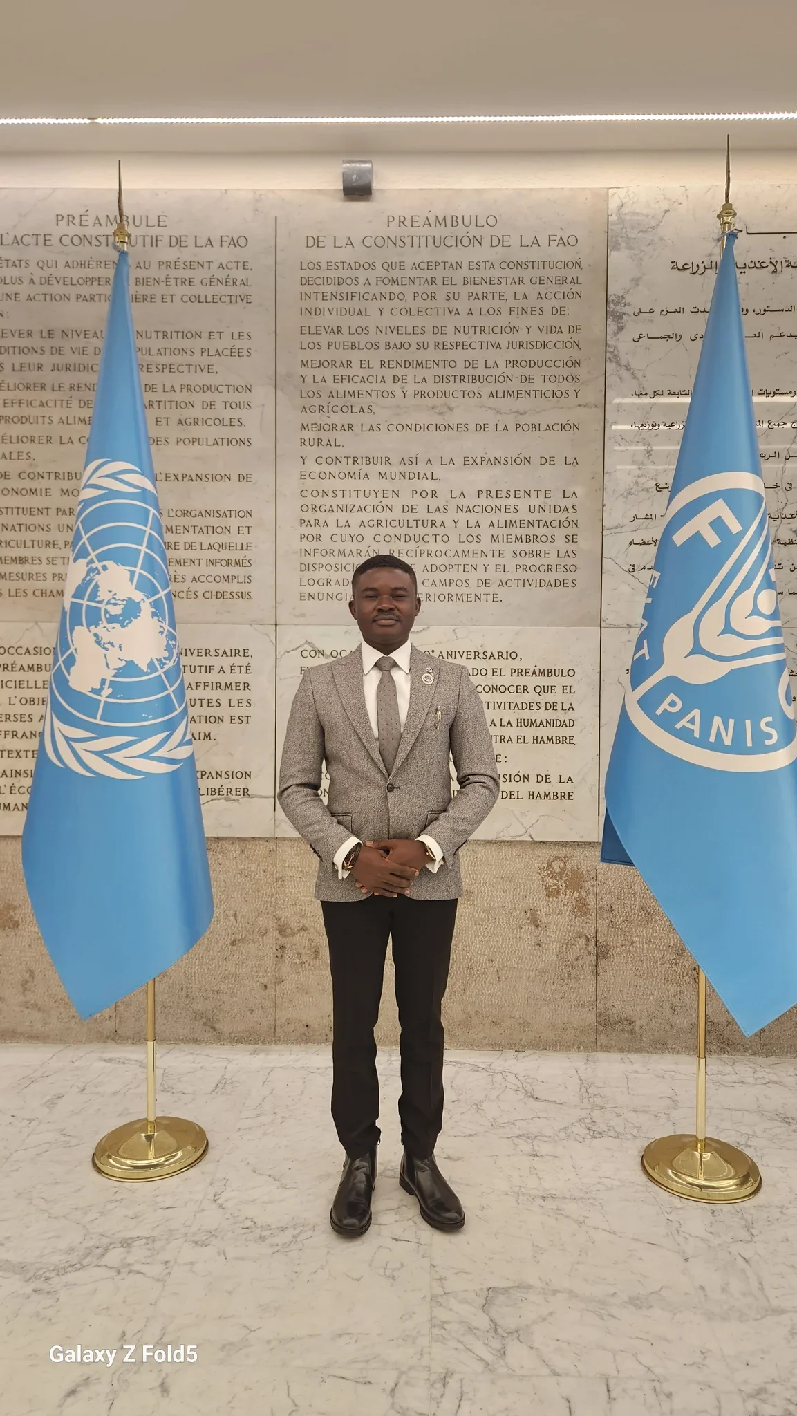 Full-length portrait of Dr. Alabi in a grey tailored suit and tie standing centrally between UN flags in the FAO preamble hall.