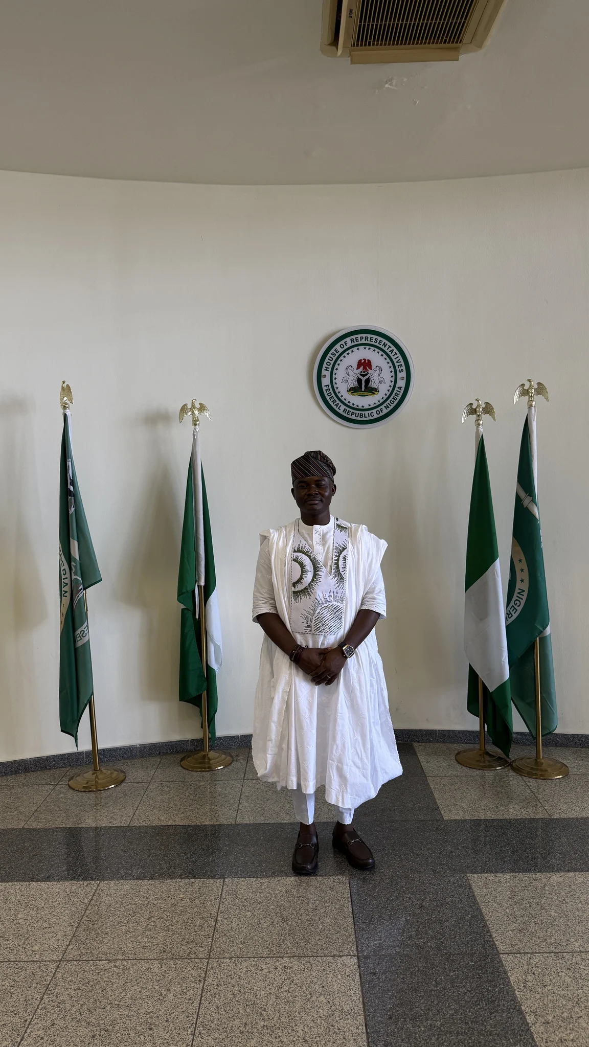 Full-length portrait of Dr. Alabi in white Agbada standing between four Nigerian national flags directly under the House of Representatives seal.