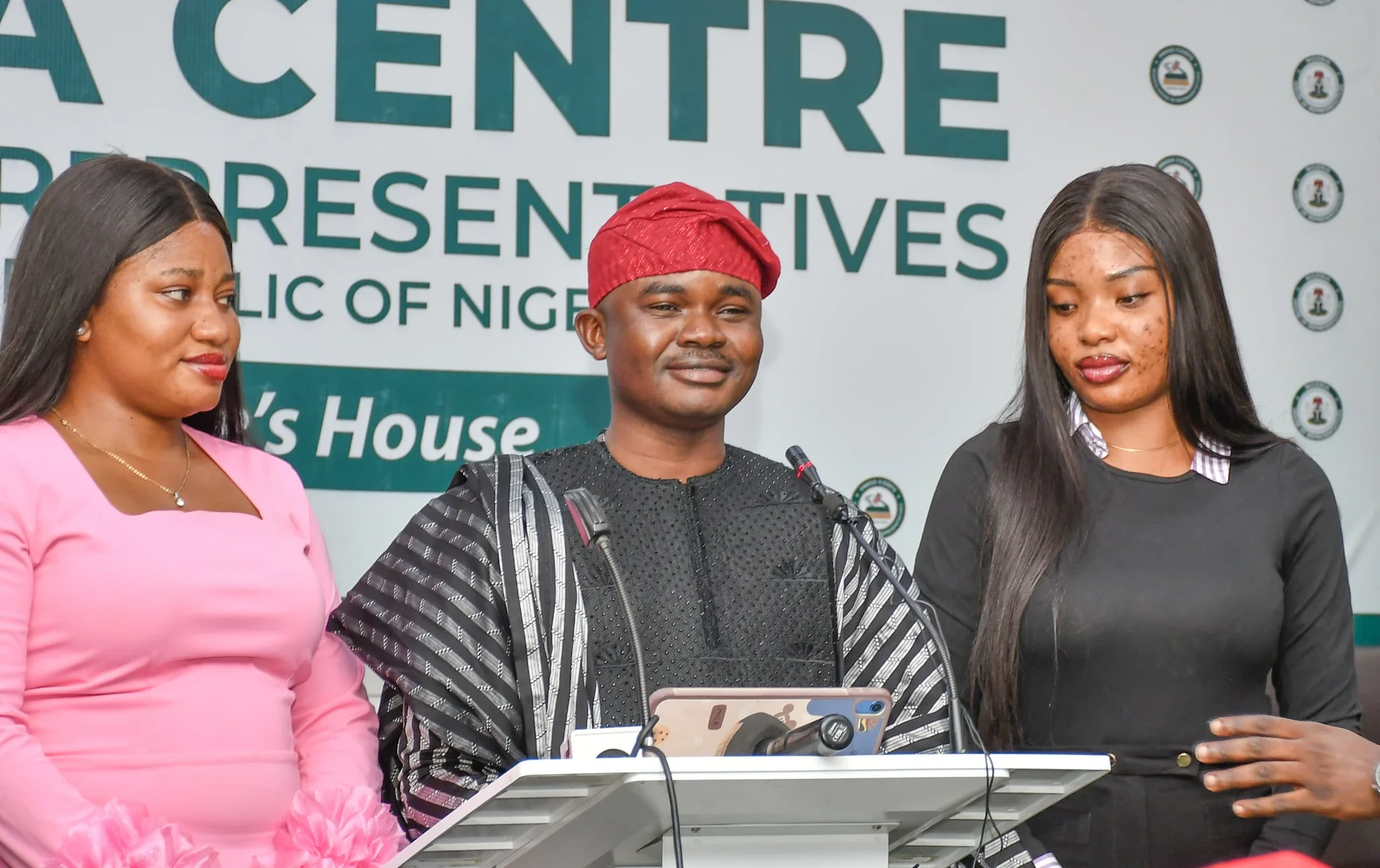 Dr. Segun Alabi wearing a traditional striped Agbada and red cap, speaking at a podium inside the Media Centre at the House of Representatives ("The People's House") flanked by two female assistants.