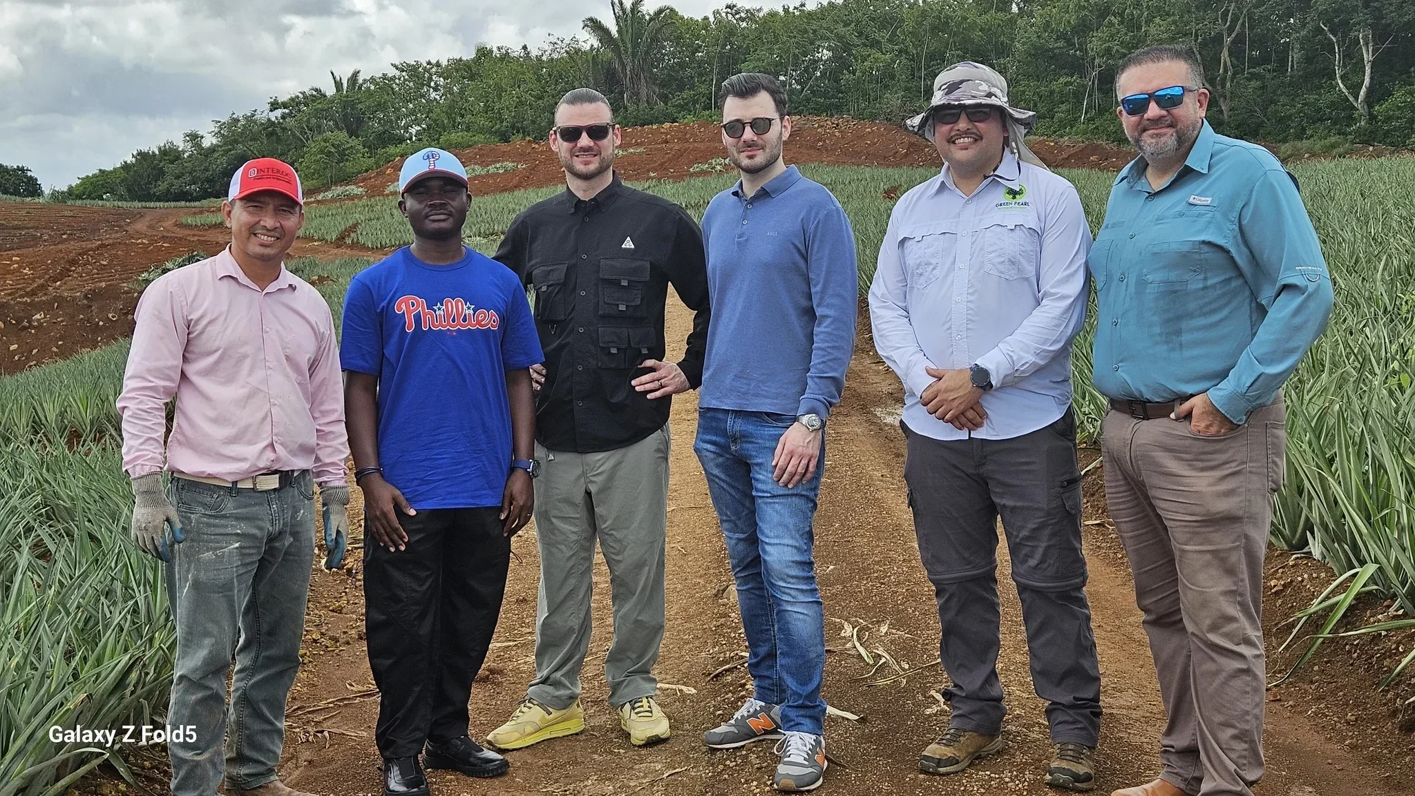 Dr. Alabi in a casual Phillies t-shirt and cap standing on the dirt path of a pineapple estate with a delegation of international partners and farm directors.