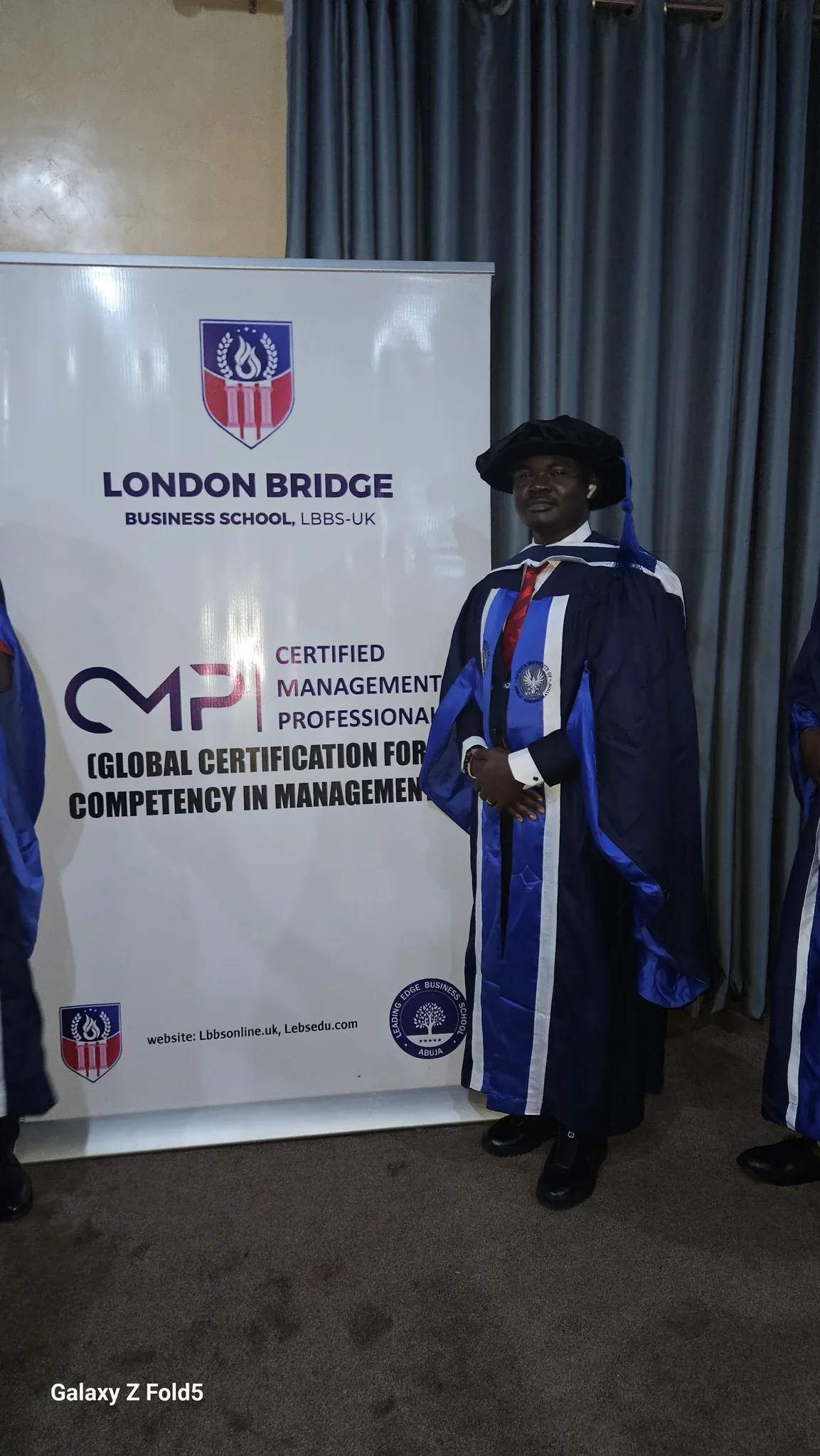 Full-length photo of Dr. Alabi in a dark blue academic gown standing beside the London Bridge Business School / Certified Management Professional banner.