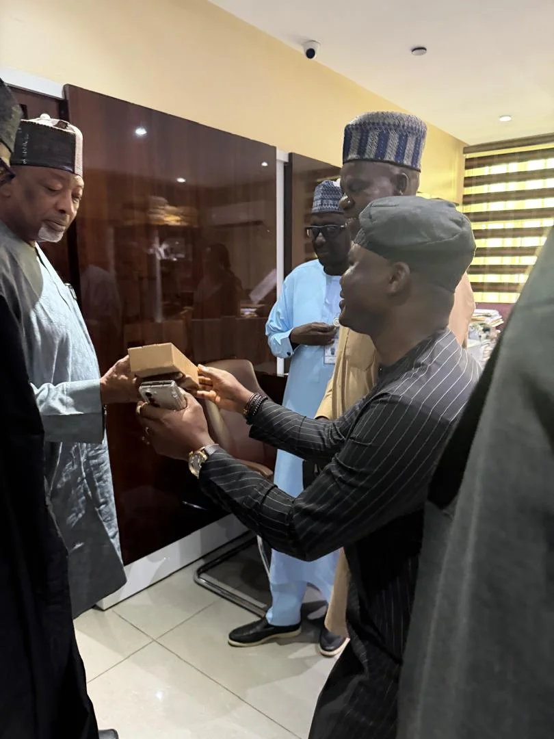 Side angle of Dr. Alabi handing an official wooden award/presentation box to the Minister of Agriculture, Senator Abubakar Kyari, surrounded by aides.