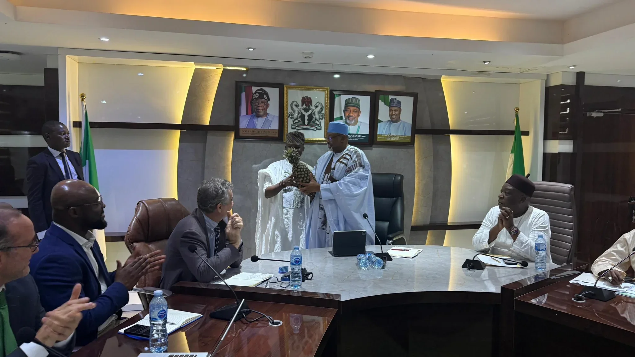 Wide boardroom photo showing Dr. Alabi in white Agbada presenting a large pineapple directly to Minister Abubakar Kyari at the head of an executive conference table, surrounded by seated delegates and international advisors.