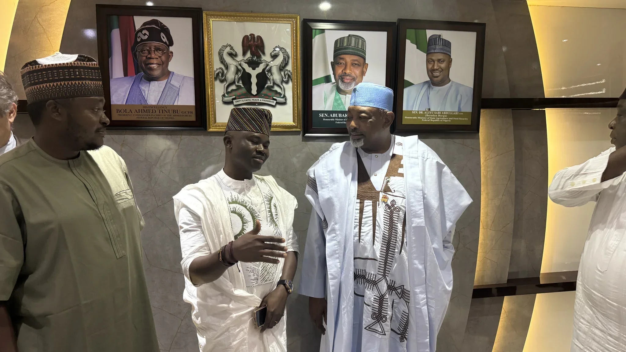 Dr. Segun Alabi in white traditional Agbada gesturing actively while consulting directly with Minister of Agriculture Senator Abubakar Kyari and senior aides under federal presidential portraits.
