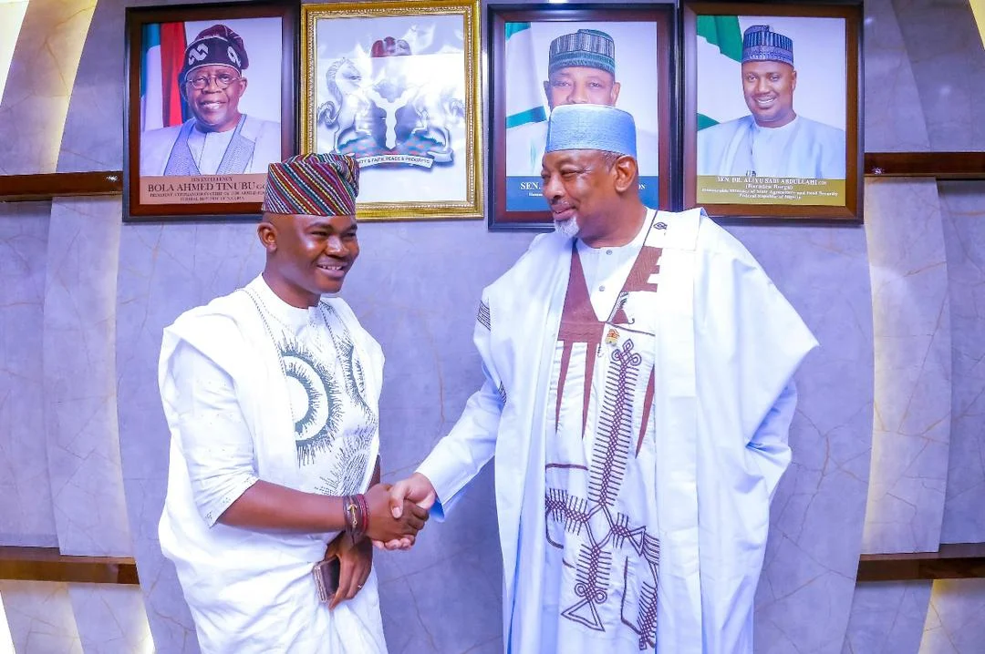 Dr. Alabi in white Agbada shaking hands warmly with Minister of Agriculture Senator Abubakar Kyari beneath official presidential portraits.