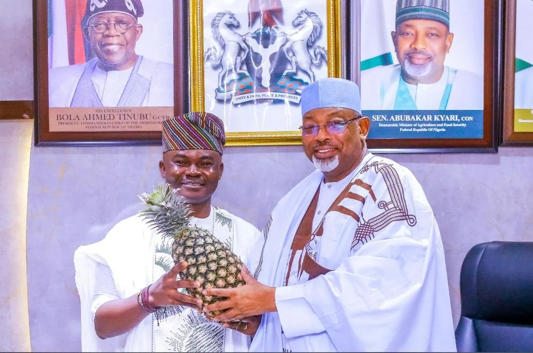 Dr. Alabi alongside Minister Abubakar Kyari, proudly presenting a premium harvested pineapple in front of presidential and ministerial portraits.
