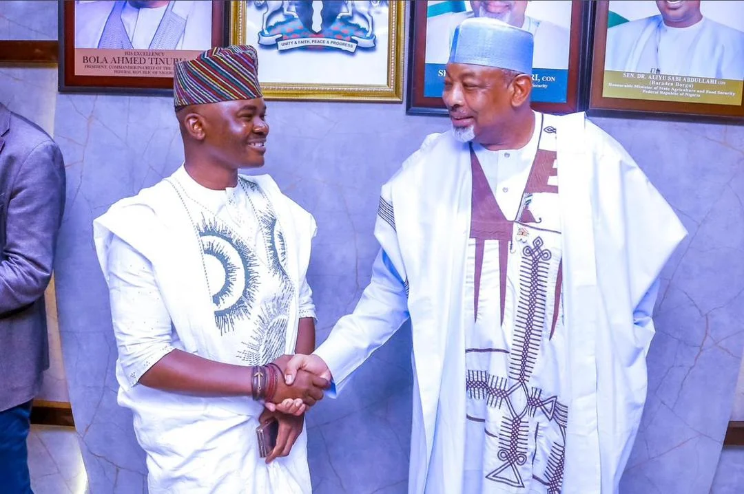 Dr. Alabi in a white Agbada sharing a warm smile and handshake with Minister of Agriculture Senator Abubakar Kyari under presidential portraits.