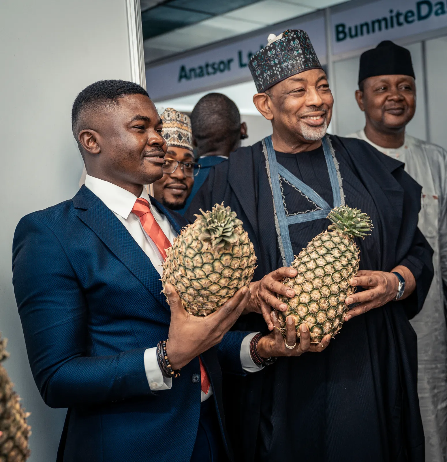 Dr. Alabi, dressed in a sharp navy blue suit and red tie, stands alongside the Minister of Agriculture and Food Security, Senator Abubakar Kyari. Both are smiling as they hold up large, ripe pineapples at an indoor exhibition or trade fair.