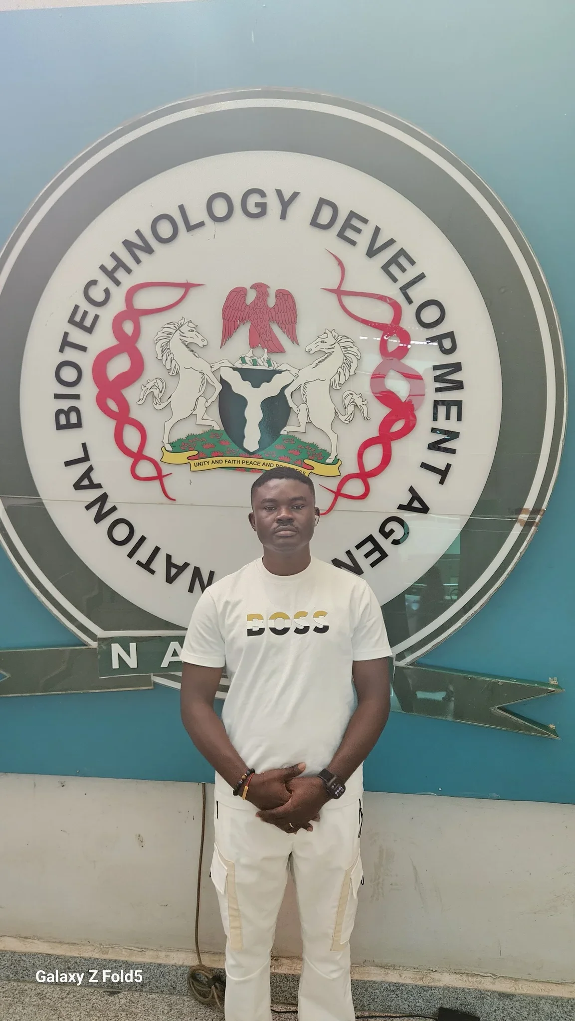 Full-length portrait of Dr. Alabi standing in front of the National Biotechnology Development Agency (NABDA) official seal.