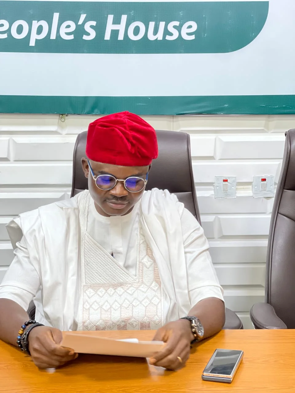 Close-up shot of Dr. Alabi in a white traditional Agbada and red cap seated attentively at an executive desk inside "The People's House" reviewing documents.