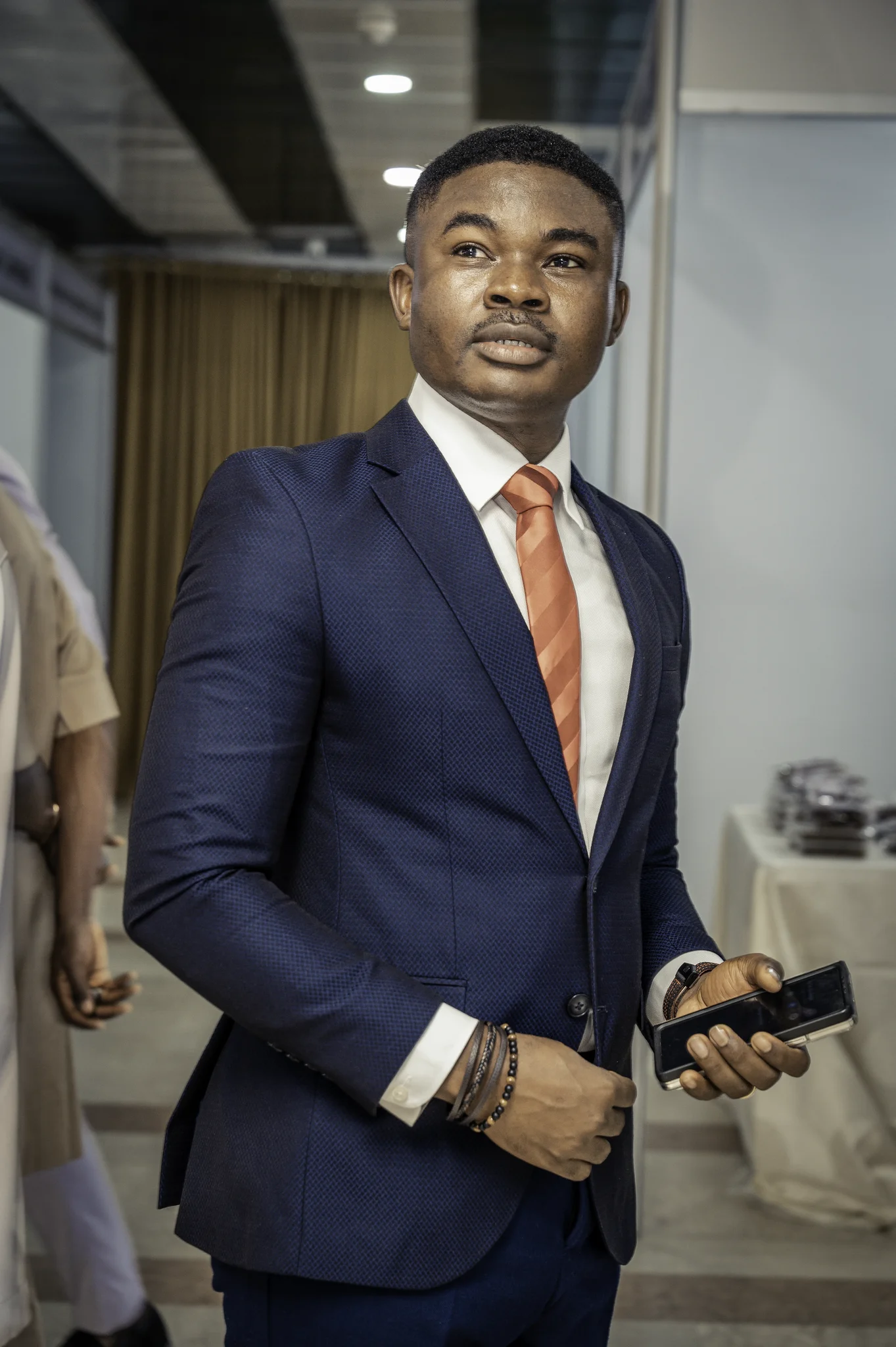 Crisp, high-resolution portrait of Dr. Alabi in a fitted navy suit and orange tie looking thoughtfully off-camera while holding a smartphone.