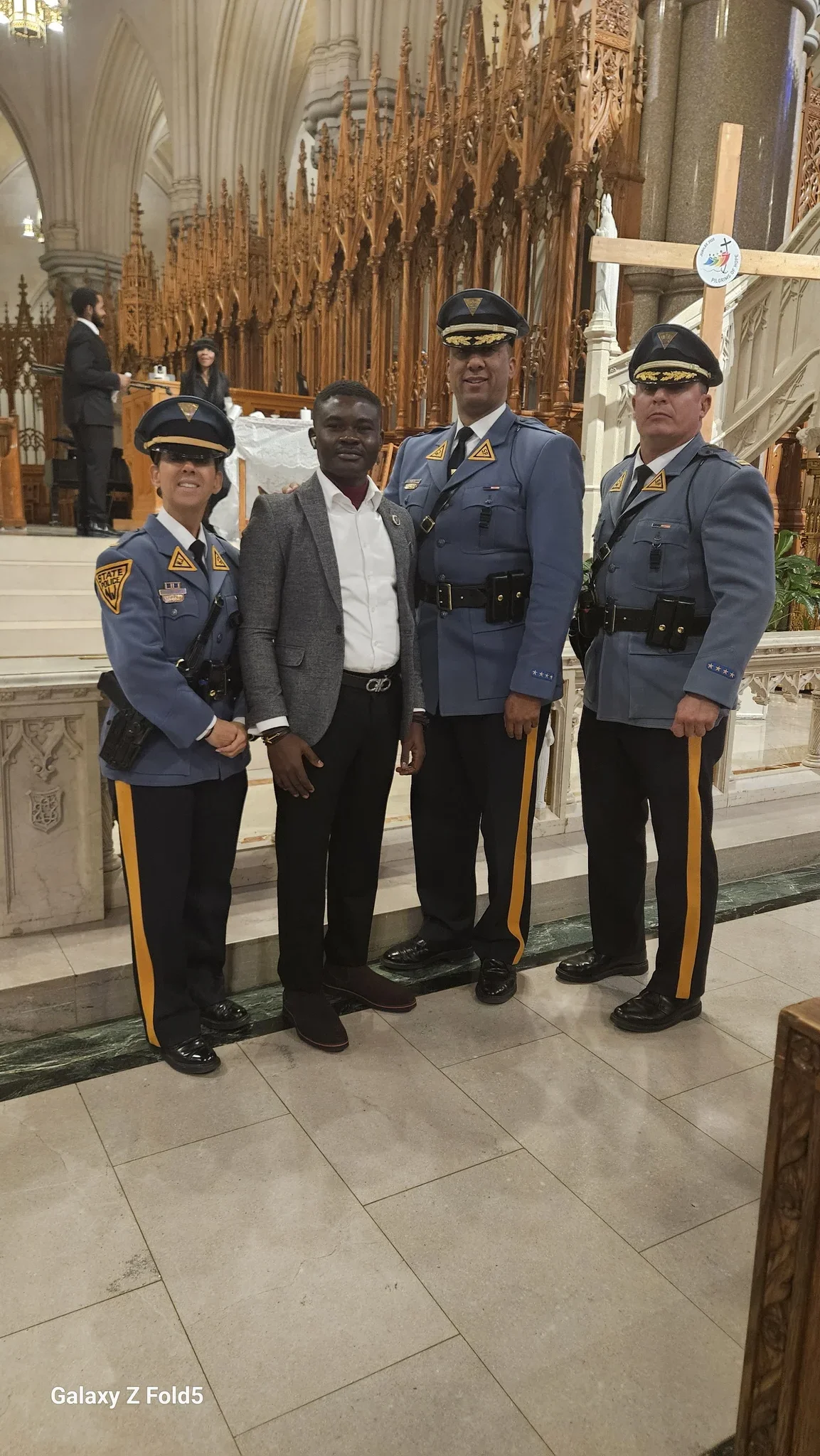 Dr. Segun Alabi in a grey blazer and black trousers standing alongside three New Jersey State Police officers in full dress uniform inside a cathedral setting.