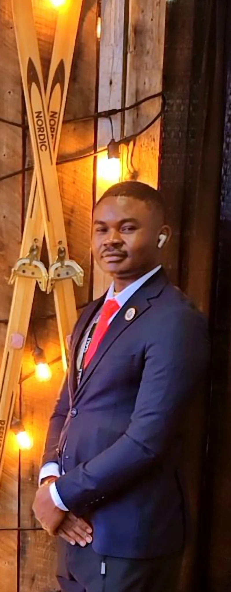 Dr. Segun Alabi in a fitted navy suit and red tie posing in front of a rustic wooden wall with crossed wooden "Nordic" skis and warm ambient string lighting.