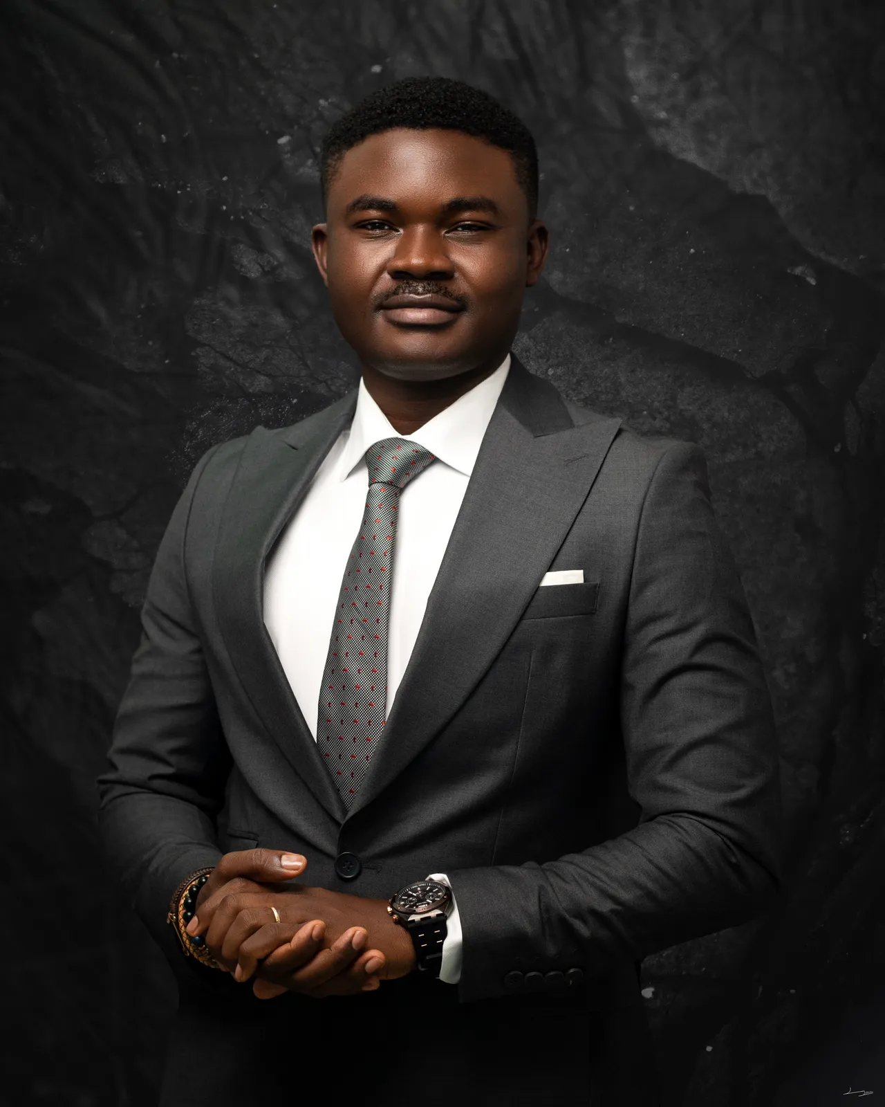 Formal, front-facing studio portrait of Dr. Alabi in a grey suit with clasped hands.