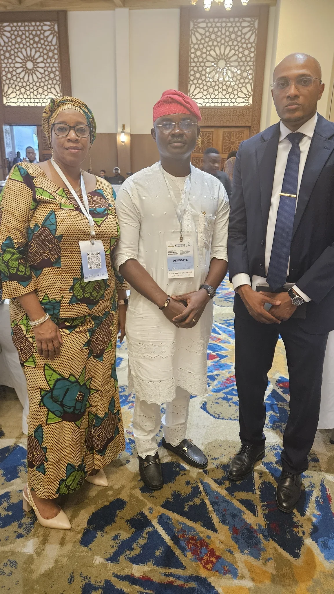 Full-length photo of Dr. Alabi flanked by a female delegate in yellow patterned lace and a male executive in a navy suit inside an ornate conference room.