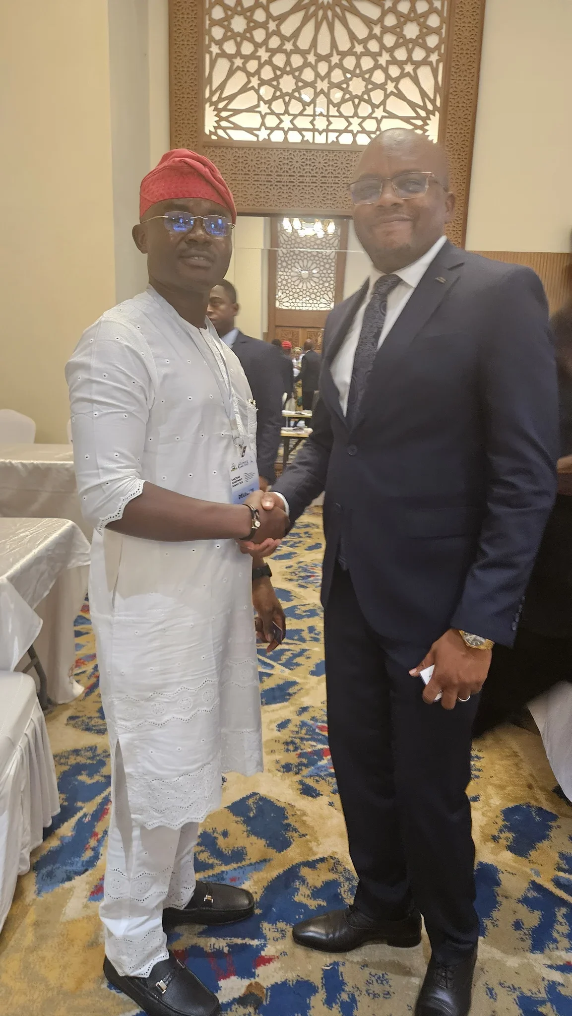 Dr. Alabi shaking hands with a corporate executive in a navy suit inside an ornate conference venue.