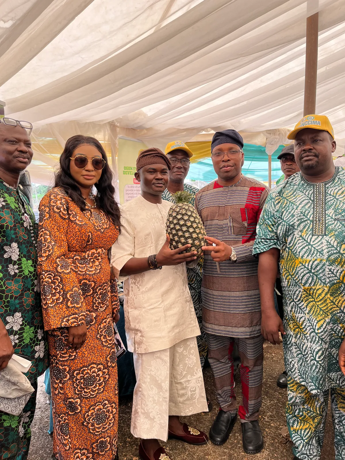 Dr. Alabi I. Oluwasegun standing in the center of a group under an outdoor canopy event, holding a large premium pineapple. He is accompanied by colleagues and delegates, including a woman in an orange patterned gown and men wearing traditional attire and caps.