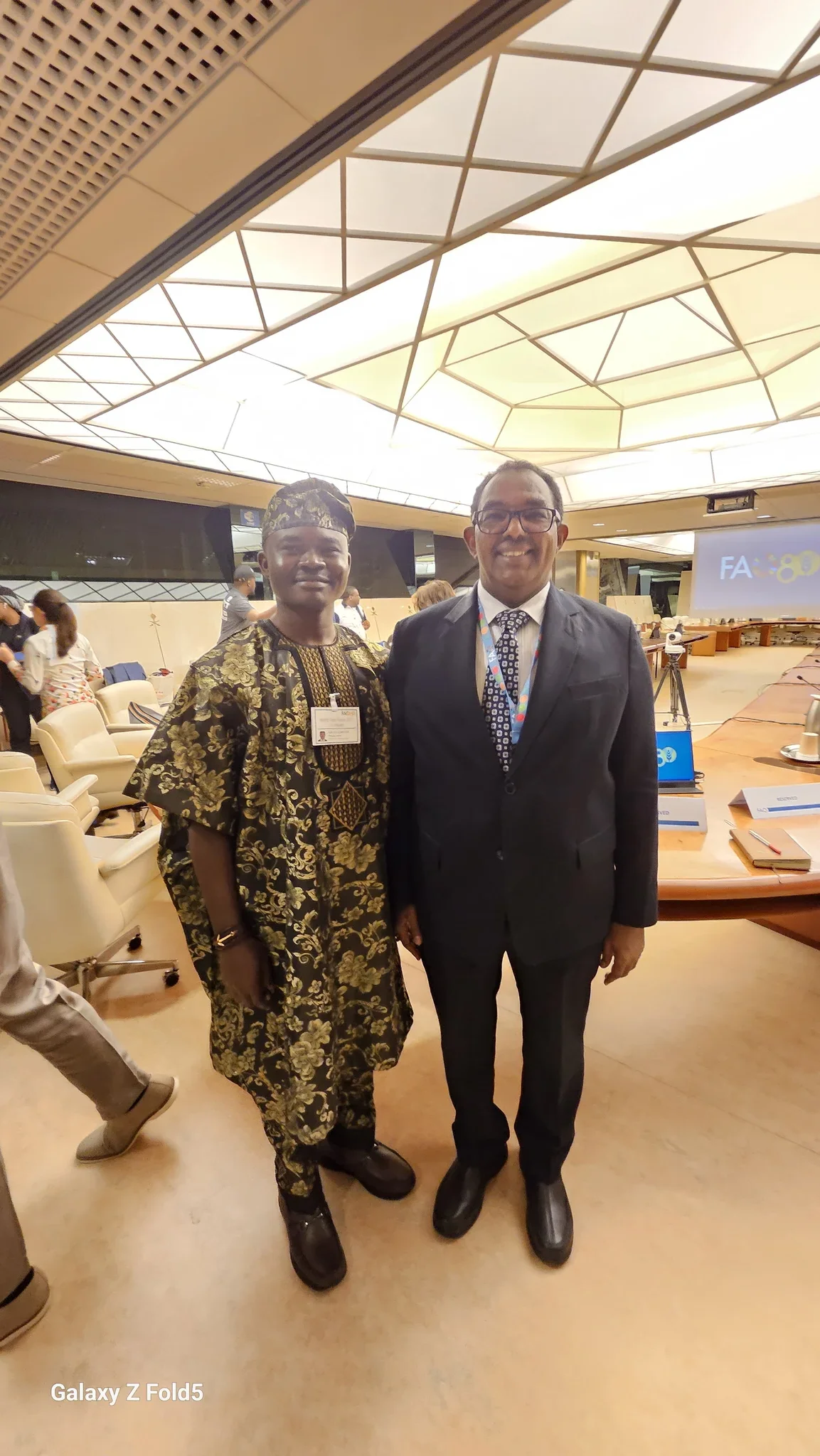 Full-length photo of Dr. Alabi in a traditional gold-patterned Agbada standing beside an international delegate inside the FAO assembly hall in Rome.