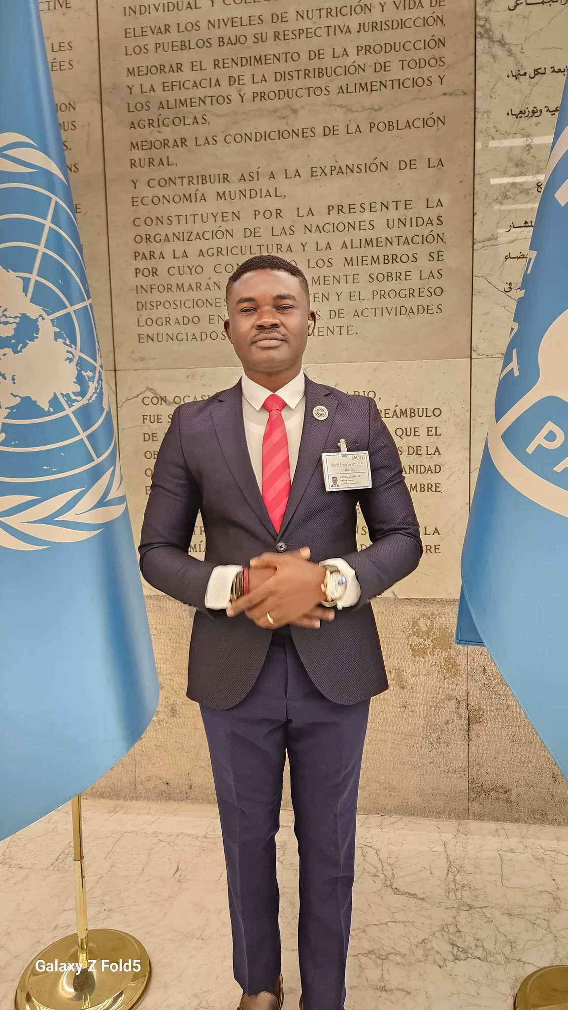 Medium-close-up portrait of Dr. Alabi in a navy suit and red tie standing between UN flags in front of the FAO constitutional wall.