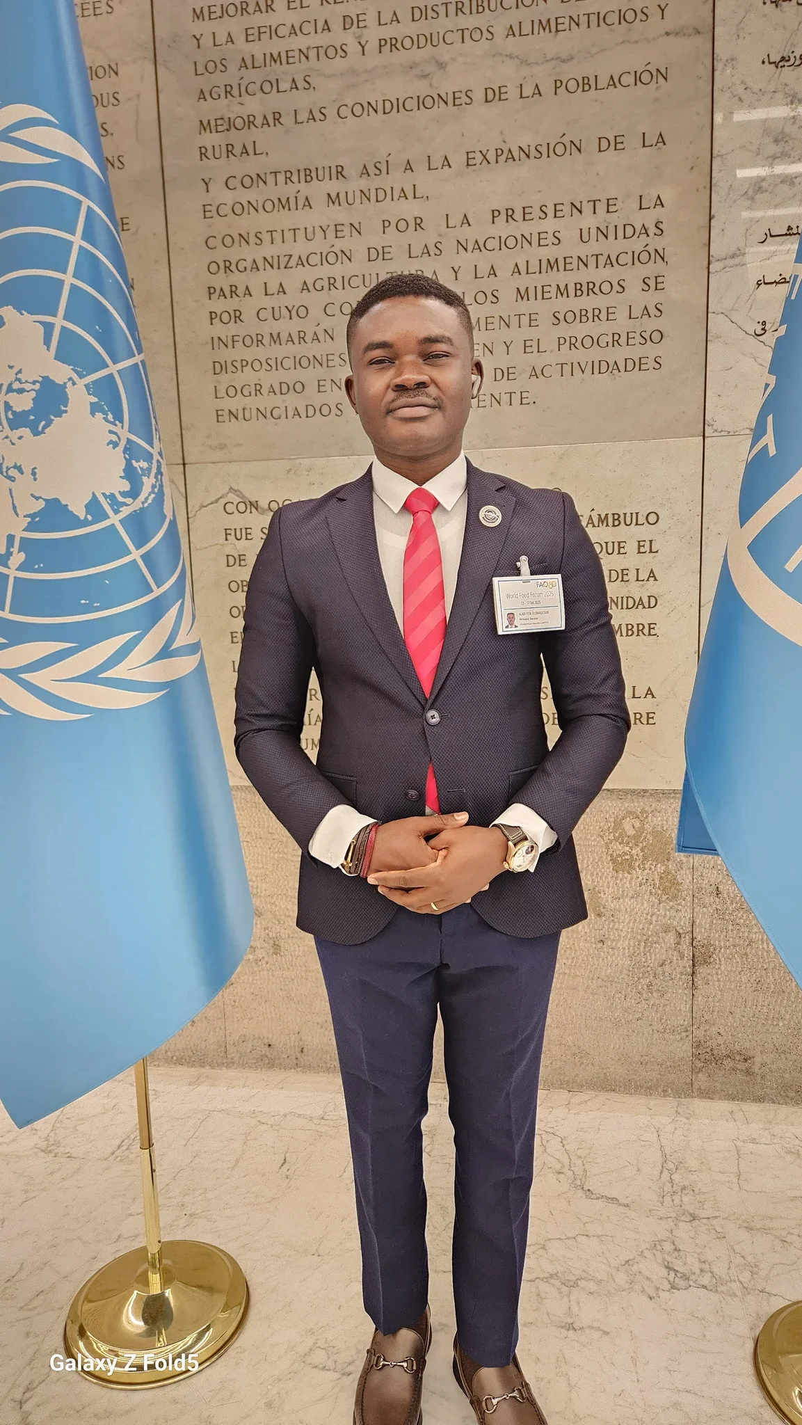 Full-length formal portrait of Dr. Alabi in a navy suit and red tie standing centrally between UN flags at the FAO Headquarters in Rome.