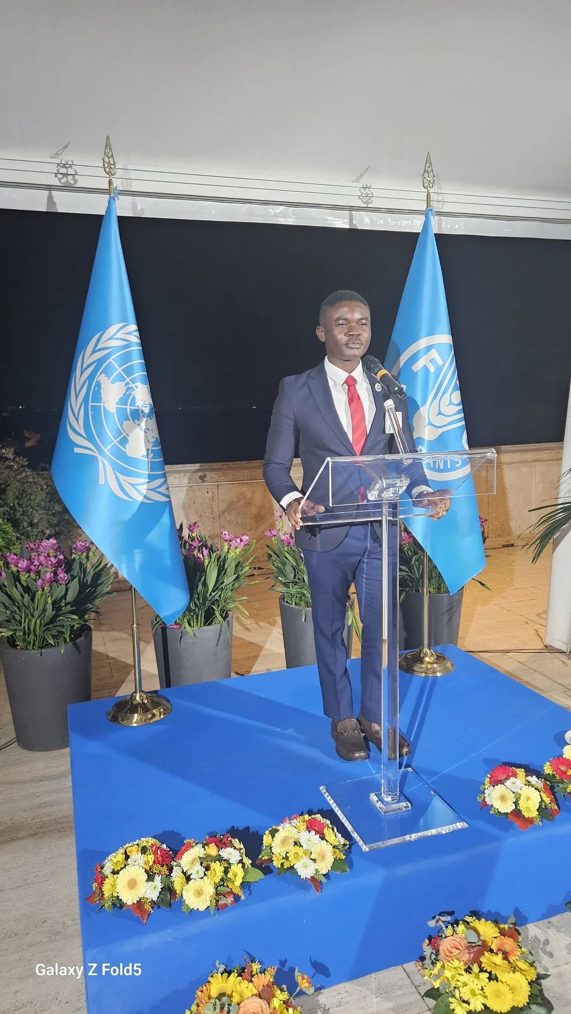 Full-length shot of Dr. Alabi delivering a keynote address from the glass podium flanked by UN flags at the FAO Headquarters stage in Rome.