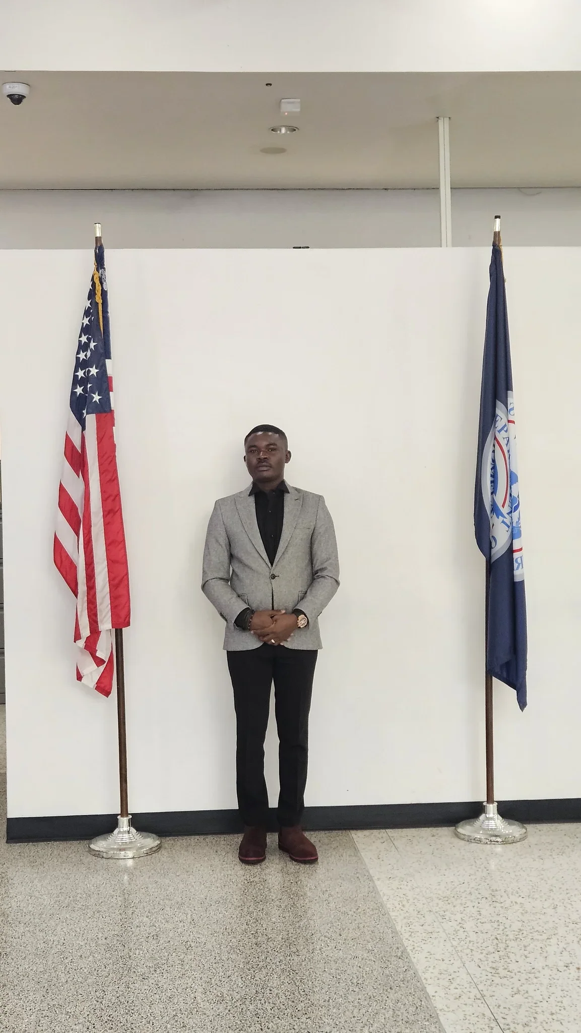 Full-length portrait of Dr. Alabi standing formally between the US flag and an official state flag against a clean white backdrop.