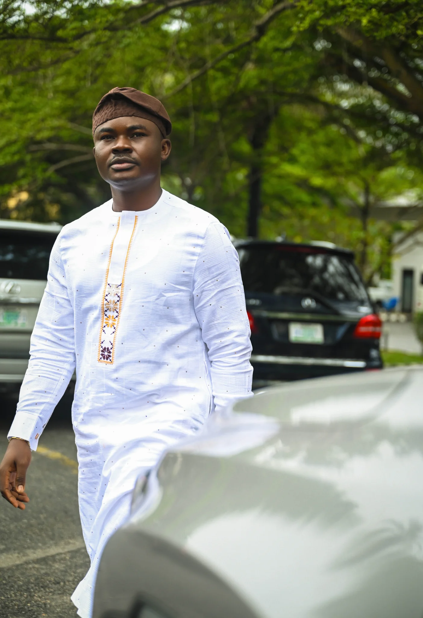Full-length outdoor candid shot of Dr. Alabi dressed in an embroidered white outfit and brown cap, walking gracefully past parked cars.